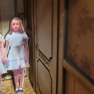 Puede incluir: Un modelo de madera de un pasillo con una puerta etiquetada con "233" y "REDRUM" en letras invertidas. Dos figuras de ni&ntilde;as con vestidos azules se encuentran en el pasillo.