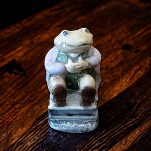 May include: A ceramic frog figurine sitting on a small platform. The frog is wearing a purple jacket and green pants. The frog has a light brown and white color scheme.