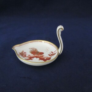 May include: A white porcelain swan-shaped dish with a red and gold design. The dish has a small handle in the shape of a swan's neck.