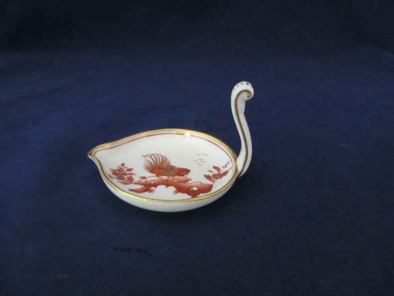 May include: A white porcelain swan-shaped dish with a red and gold design. The dish has a small handle in the shape of a swan's neck.