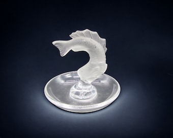 May include: A clear glass ring holder shaped like a fish with a frosted finish. The fish is curled up with its tail in the air and its head resting on a small, round dish.