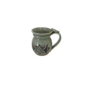 May include: A handmade ceramic mug with a pale green glaze. The mug has a rounded body and a handle with a small, decorative element. The front of the mug is decorated with geometric shapes in red, black, and white.