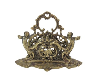 May include: A gold metal napkin holder with an ornate design featuring two cherubs and a decorative scrollwork pattern.