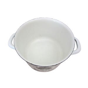 K&ouml;nnte beinhalten: Wei&szlig;er Emaille-Topf mit zwei Griffen. Der Topf hat einen schwarzen Rand und ein dekoratives Muster um den Boden. Der Innenraum ist wei&szlig; und die Griffe sind gebogen. Ein K&uuml;chenartikel.