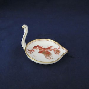 May include: A white porcelain swan-shaped dish with a red and gold floral design. The dish has a handle that resembles a swan's neck.