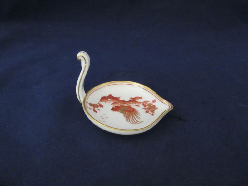May include: A white porcelain swan-shaped dish with a red and gold floral design. The dish has a handle that resembles a swan's neck.