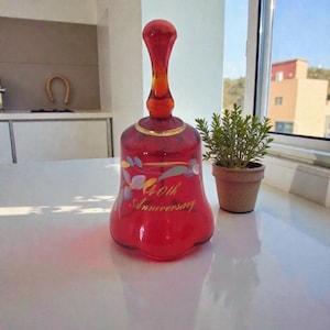 May include: A red glass bell with a long handle and a gold rim. The bell features the words "40th Anniversary" and floral designs. A small potted plant is next to it.