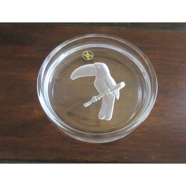 Puede incluir: Un posavasos de cristal transparente con un relieve de un tuc&aacute;n blanco posado en una rama. El posavasos tiene un emblema dorado y es redondo. Descansa sobre una superficie de madera marr&oacute;n oscuro.