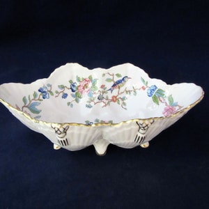 May include: A white porcelain bowl with a scalloped edge and three feet. The bowl is decorated with a floral pattern and a blue bird on a branch.