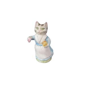 May include: A white and brown cat figurine wearing a blue dress and holding a yellow object. The figurine is standing on a green base.