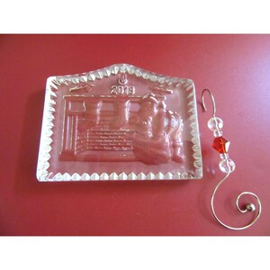 May include: A clear glass rectangular ornament featuring a relief design of Santa Claus by a fireplace with stockings and the year 2013 engraved at the top. It includes a gold-toned metal hook adorned with two clear beads and one red faceted bead.
