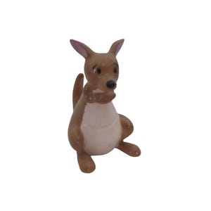 May include: A small, brown kangaroo figurine with a white belly and pink-tipped ears. The kangaroo is in a seated position, with its tail curled behind it. The figurine has a detailed, cartoon-like appearance.