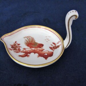 May include: A white porcelain dish with a gold rim and a red rooster design. The dish has a long, curved handle.