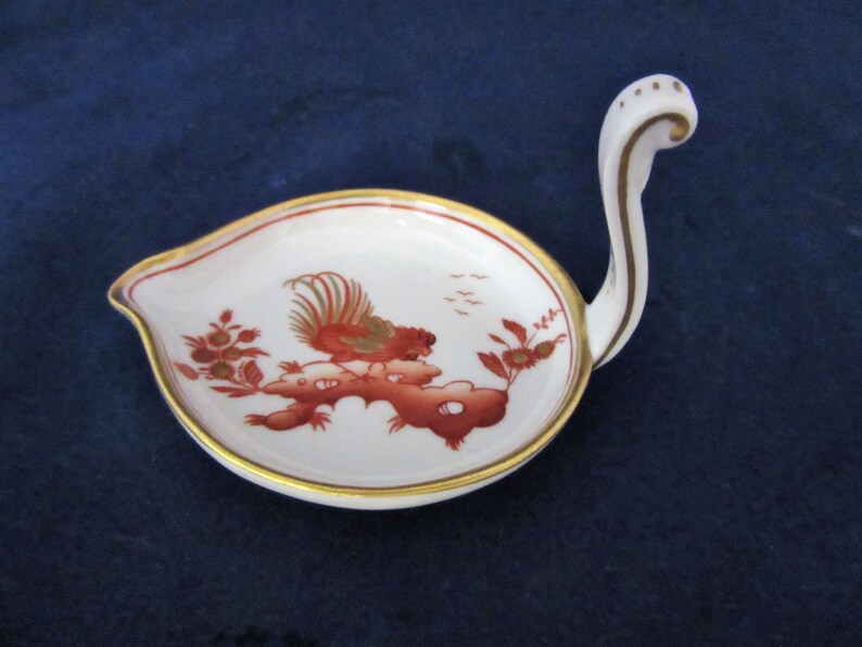 May include: A white porcelain dish with a gold rim and a red rooster design. The dish has a long, curved handle.