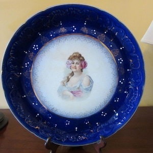 May include: A blue and white porcelain plate with a raised rim and a gold trim. The center of the plate features a painted image of a woman with flowers in her hair.