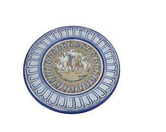 May include: Round ceramic plate with a detailed illustration of two figures on horseback in a landscape scene. The plate features a blue and white border with a repeating arched design. The central image has yellow, green, and brown tones.
