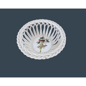 May include: A white porcelain basket with a woven design and a braided rim. The center features a circular design with a painted image of two birds perched on a branch. The birds are brown and green.