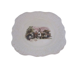 May include: A white decorative plate with a scalloped edge and embossed floral design. The center features a painted scene of a village with buildings, trees, and a pink sky. The plate is a vintage piece, likely for display.