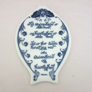 May include: A white ceramic plaque with blue floral designs and a Danish proverb in blue script: "At smörrebrod er ikke mad, og hierlighed er ikke had. Det er for tiden hvad jeg ved om smörrebrod oy hierlighed." The plaque is signed "WESSEL".