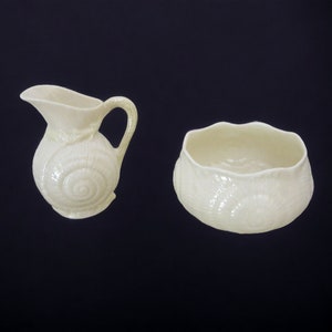 May include: A cream-colored ceramic creamer and sugar bowl set with a shell design. The creamer has a handle and a spout. The sugar bowl is round and has a scalloped edge.