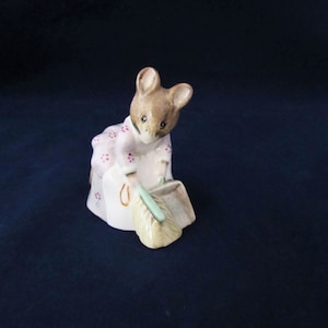 May include: A small ceramic figurine of a mouse wearing a pink dress with white flowers and holding a broom and dustpan. The mouse is brown and white with a pink nose.