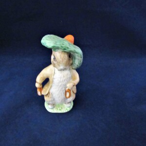 May include: A ceramic figurine of Peter Rabbit wearing a green hat with an orange band. He is holding a brown stick in his right hand and a brown stick in his left hand. He is standing on a green base.