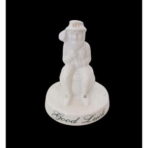 May include: A white porcelain figurine of a seated leprechaun wearing a hat, resting on a barrel-shaped base. The base is round and has the words "Good Luck" in cursive. Gold accents are on the hat and shoes. The figurine is on a black background.