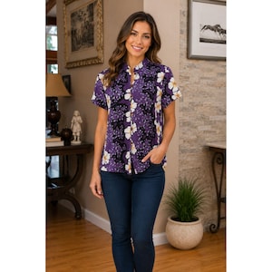 May include: A purple short-sleeve shirt with a floral pattern of white and yellow flowers. The shirt has a mandarin collar and is paired with dark blue jeans. The woman is standing in a well-lit room with artwork on the walls.