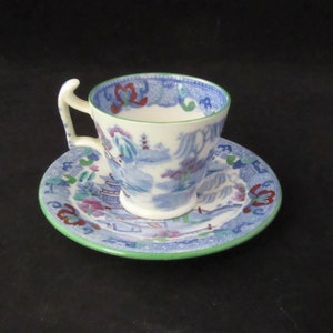 May include: A blue and white teacup and saucer with a green rim and a floral design. The cup has a unique handle.