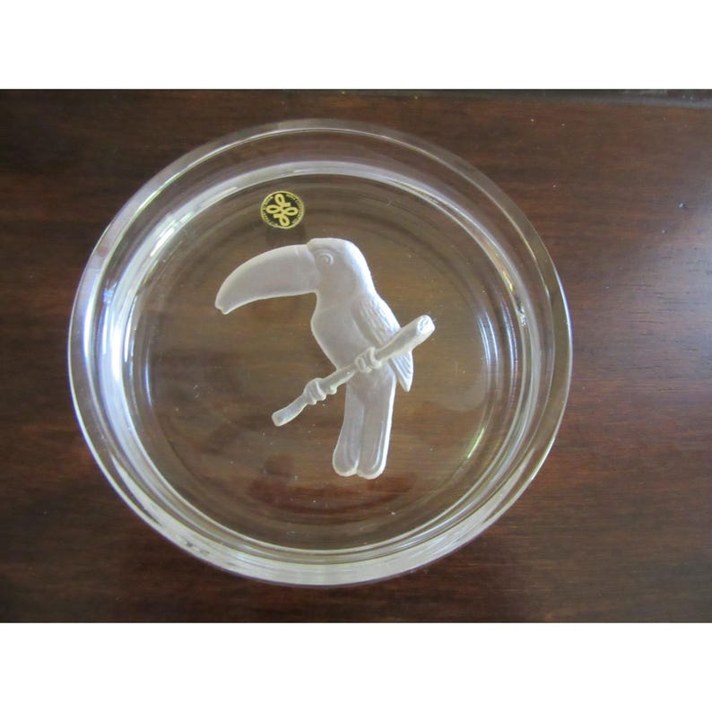Puede incluir: Un posavasos de vidrio transparente con un dise&ntilde;o de tuc&aacute;n esmerilado en una rama. El posavasos tiene forma circular y un borde ligeramente elevado. Un peque&ntilde;o emblema dorado es visible en la parte superior.