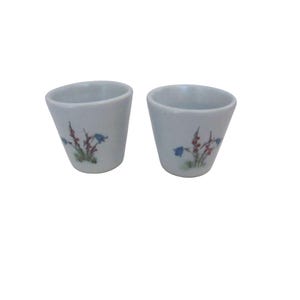 May include: Two small, white ceramic cups with a slightly flared shape. Each cup features a delicate floral design with blue bellflowers and red accents. The cups are likely for decorative use or for serving small portions.