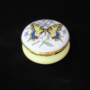 May include: A small, round, yellow trinket box with a white lid. The lid features a painted image of a yellow and black butterfly with blue and red accents, perched on a green stem with purple flowers.