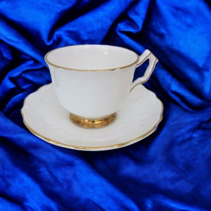 May include: A white teacup and saucer with a gold trim. The teacup and saucer are on a blue satin fabric.