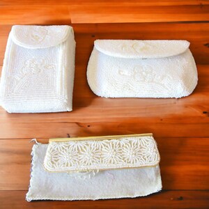 May include: Three white beaded purses with different designs. The purses are on a wooden surface.