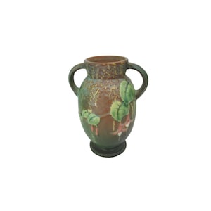 May include: A brown and green ceramic vase with two handles. The vase features a textured surface and is decorated with green leaves and pink floral accents. The vase has a dark green base and a brown rim.