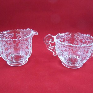 May include: A vintage glass creamer and sugar bowl set with a delicate etched floral pattern. The set is clear glass with a scalloped edge and ornate handles.