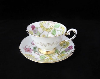 May include: A white teacup and saucer with a floral pattern in shades of pink, yellow, and green. The cup has a gold rim and a gold handle. The saucer has a gold rim.