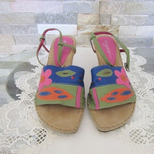May include: A pair of colorful sandals with a floral design. The sandals feature a tan sole, a blue, green, and orange upper with a pink flower, and green and red straps. The sandals are displayed on a white surface.