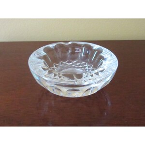 May include: A clear, round ashtray with a cut glass design. The ashtray has a series of indentations around the rim for resting cigarettes. The interior features a starburst pattern. The ashtray is sitting on a dark brown surface.