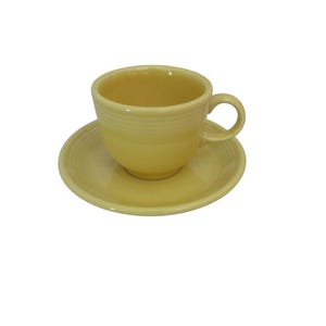 May include: A pale yellow teacup and saucer set. The cup has a rounded shape with a handle and is sitting on a matching saucer. The set is made of ceramic and has a smooth, glossy finish. The design is simple and classic.