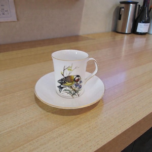 May include: A white porcelain teacup and saucer set featuring a detailed illustration of a goldfinch perched on a branch with blue flowers. The cup and saucer are finished with a thin gold rim along the edges and a curved handle on the cup.