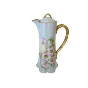 May include: A vintage porcelain creamer with a delicate floral design. The creamer is white with a light blue glaze and features pink and green flowers. The handle is gold-trimmed.