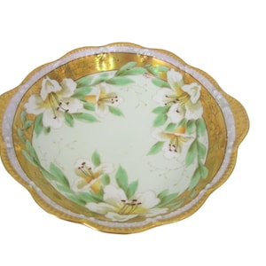 May include: A round, scalloped-edge serving dish with a pale green interior. The dish features a gold rim and is decorated with white and yellow lily flowers and green leaves. The rim has a gold and white border.