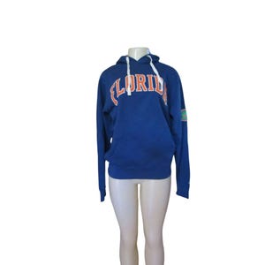 May include: A blue hooded sweatshirt with the word "FLORIDA" in orange and white lettering across the chest. The sweatshirt has a white drawstring and a small patch on the left sleeve. The garment is displayed on a mannequin.
