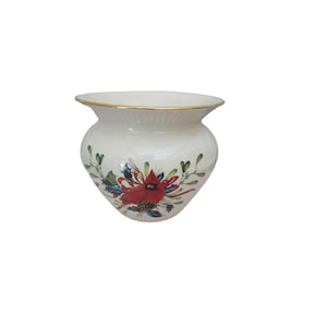 May include: A white ceramic vase with a gold rim and a rounded body. The vase is decorated with a red cardinal bird and green and blue foliage. The vase is a decorative item.