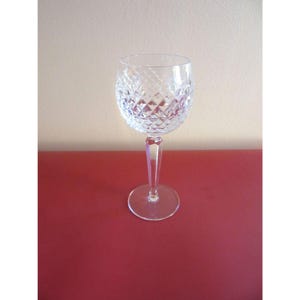 May include: A clear crystal wine glass with a diamond-cut pattern on the bowl. The glass has a long, faceted stem and a round base. The glass is set against a red surface.