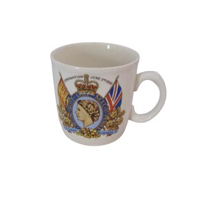 May include: White ceramic mug featuring a commemorative design. The mug has a handle and displays a royal emblem with the text "CORONATION JUNE 2 1953" and flags. The design includes a portrait of a woman.