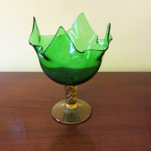 May include: A green glass bowl with a ruffled edge and a twisted amber stem. The bowl is on a wooden surface.