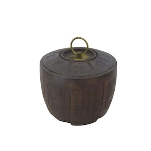 May include: A dark brown, cylindrical container with a lid. The lid has a brass ring handle and is decorated with etched symbols. The container's sides feature etched designs and text, suggesting an Asian cultural influence.