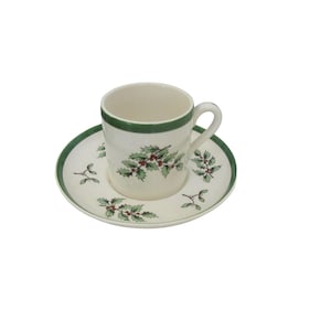 May include: A white ceramic teacup and saucer with a green rim and a holly leaf design.